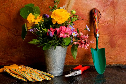 A garden scene featuring a concrete planting pot filled with a variety of colorful flowers, including yellow, pink, and purple blooms, along with lush green leaves. The pot is placed on a dark surface against a warm, textured orange-brown wall. To the right of the pot, there is a small green-handled garden trowel with a wooden handle, leaning against the wall. Nearby, a pair of pruning shears with red handles and a pair of yellow gardening gloves with striped patterns are also positioned on the surface, indicating garden maintenance activity. The image reflects a well-organized gardening workspace, with tools and plants ready for outdoor care, subtly conveying services offered by Garden Maintenance Marylebone in London, with hints of local context through the style and colour palette that complements outdoor garden environments. The natural lighting highlights the vibrant colours of the flowers and the textures of the tools, suggesting a bright and inviting garden setting suitable for maintenance or planting projects.